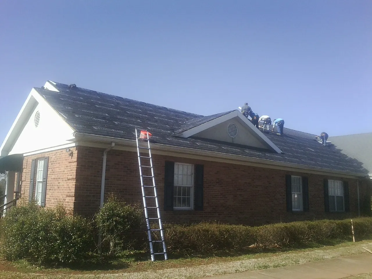 Crew tearing shingles off a brick ranch roof with a ladder set — Metal Roof Repair work by our crew in Blockton