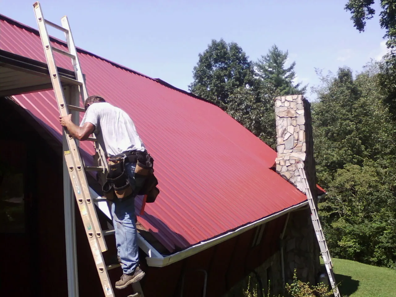 Roofer on a ladder beside a red metal roof and stone chimney — Soffit & Fascia Repair by Roofing Brothers in Blockton